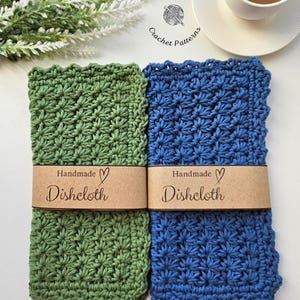 Peut inclure: Deux torchons faits à la main au crochet, un vert et un bleu, sont présentés côte à côte. Chaque tissu est enveloppé d'une bande de papier marron sur laquelle on peut lire "Handmade Dishcloth". Une tasse de café et des plantes vertes décoratives sont en arrière-plan.