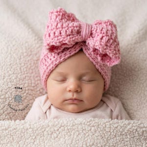 May include: A baby wearing a pink crochet bow headband. The headband is a light pink color and has a large bow on top. The baby is sleeping and is wrapped in a soft, white blanket. The text "Tina Crochet Patterns" is in the lower left corner.