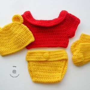 May include: A crocheted baby outfit in red and yellow. The outfit includes a hat with bear ears, a short-sleeved top, a diaper cover, and booties. The hat, diaper cover, and booties are yellow, while the top is red.