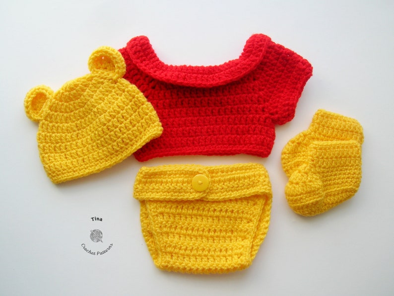 May include: A crocheted baby outfit in red and yellow. The outfit includes a hat with bear ears, a short-sleeved top, a diaper cover, and booties. The hat, diaper cover, and booties are yellow, while the top is red.