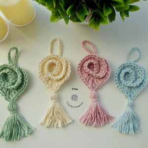 May include: Four crocheted heart-shaped ornaments with tassels in green, cream, pink, and blue. The ornaments are hanging from a white surface. Tina Crochet Patterns is written below the ornaments.
