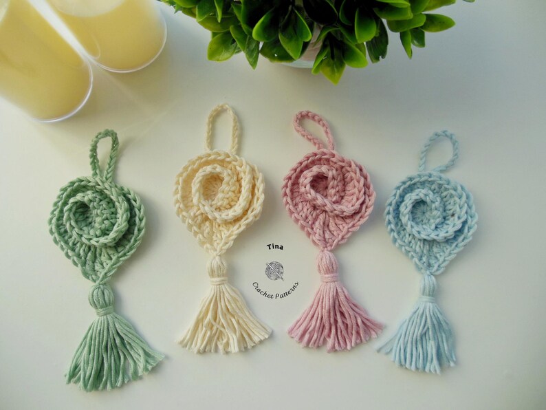 May include: Four crocheted heart-shaped ornaments with tassels in green, cream, pink, and blue. The ornaments are hanging from a white surface. Tina Crochet Patterns is written below the ornaments.