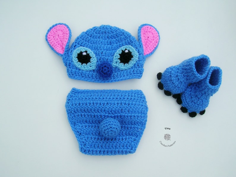 May include: A blue crocheted baby hat, diaper cover, and booties set featuring a cartoon character with pink ears, black eyes, and a blue nose. The set is made with a soft, textured yarn.