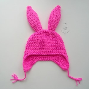 Op de afbeelding: Roze gehaakte konijnenmuts met lange oorflappen en een kwastje aan elke flap. De muts is gemaakt met een vaste steek.