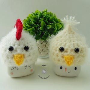 May include: Two crocheted chicken mug cozies, one with a red comb and one with a white comb, are sitting on a white surface in front of a small potted plant. The mug cozies have black eyes and yellow beaks. The text "Tina Crochet Patterns" is visible in the bottom right corner of the image.