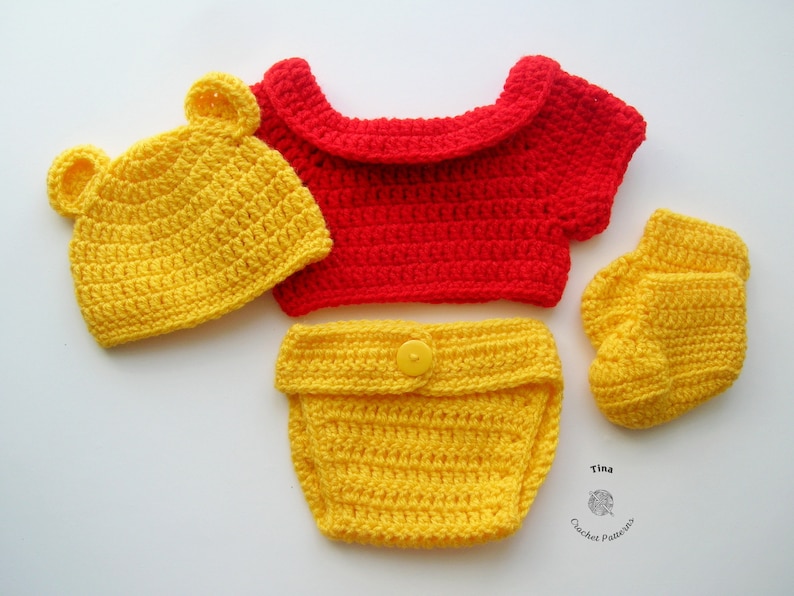May include: A crocheted baby outfit in yellow and red. The outfit includes a hat with bear ears, a short-sleeved shirt, diaper cover, and booties. The hat, diaper cover, and booties are yellow, and the shirt is red.