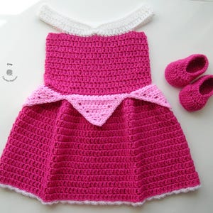 May include: A pink and white crocheted dress with a white crocheted collar and a pink crocheted crown. The dress is paired with matching pink crocheted shoes.