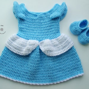 Peut inclure: Une robe de b&eacute;b&eacute; en crochet bleu clair avec des bordures blanches et des chaussures assorties en crochet bleu clair. La robe a une jupe &agrave; volants et un d&eacute;collet&eacute; arrondi.