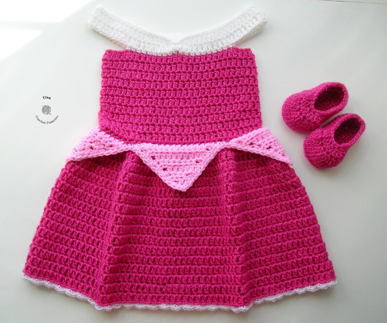 May include: A pink and white crocheted dress with a white crocheted collar and a pink crocheted crown. The dress is paired with matching pink crocheted shoes.