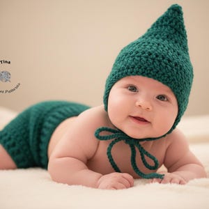 May include: A baby wearing a dark teal crochet hat with a pointed top and matching diaper cover. The hat has a tie under the chin. The baby is lying on a cream-colored blanket.