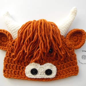 May include: A crocheted Highland cow hat in shades of orange, white, and black. The hat features a shaggy orange mane, white horns, and black button eyes. The nose is white with two black buttons. The ears are orange.