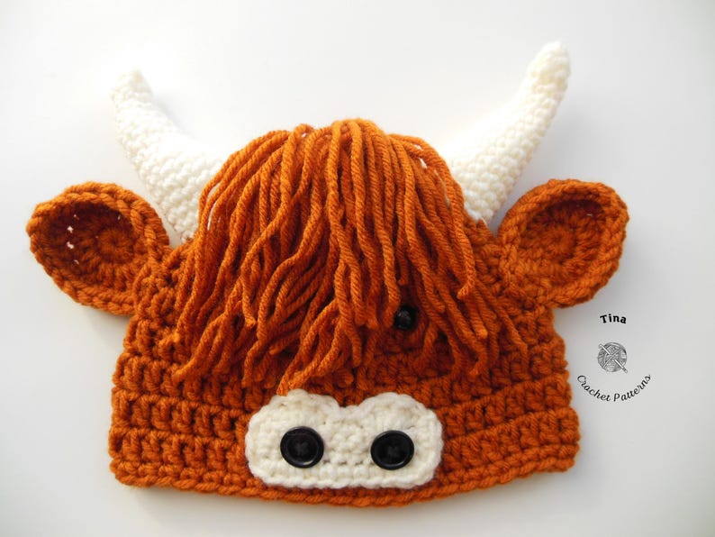 May include: A crocheted Highland cow hat in shades of orange, white, and black. The hat features a shaggy orange mane, white horns, and black button eyes. The nose is white with two black buttons. The ears are orange.