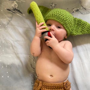 May include: A baby wearing a green crocheted Yoda hat and brown crocheted nappy, holding a green lightsaber toy.