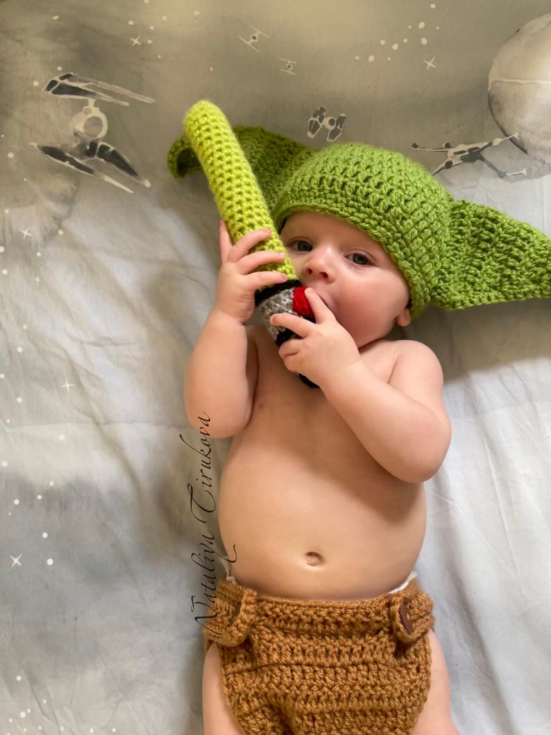 May include: A baby wearing a green crocheted Yoda hat and brown crocheted nappy, holding a green lightsaber toy.