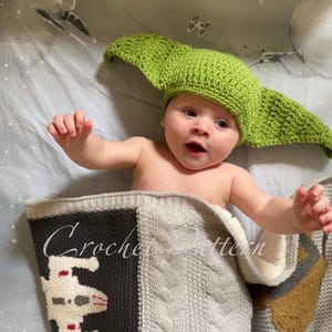 May include: A baby wearing a green crocheted Yoda hat is lying on a grey and black crocheted blanket with a Star Wars spaceship design. The blanket has the text "Crochet Pattern" on it.