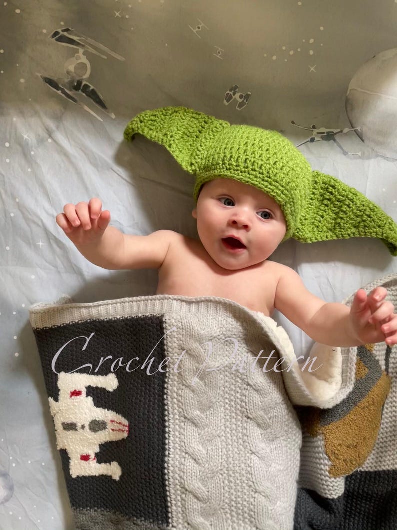 May include: A baby wearing a green crocheted Yoda hat is lying on a grey and black crocheted blanket with a Star Wars spaceship design. The blanket has the text "Crochet Pattern" on it.