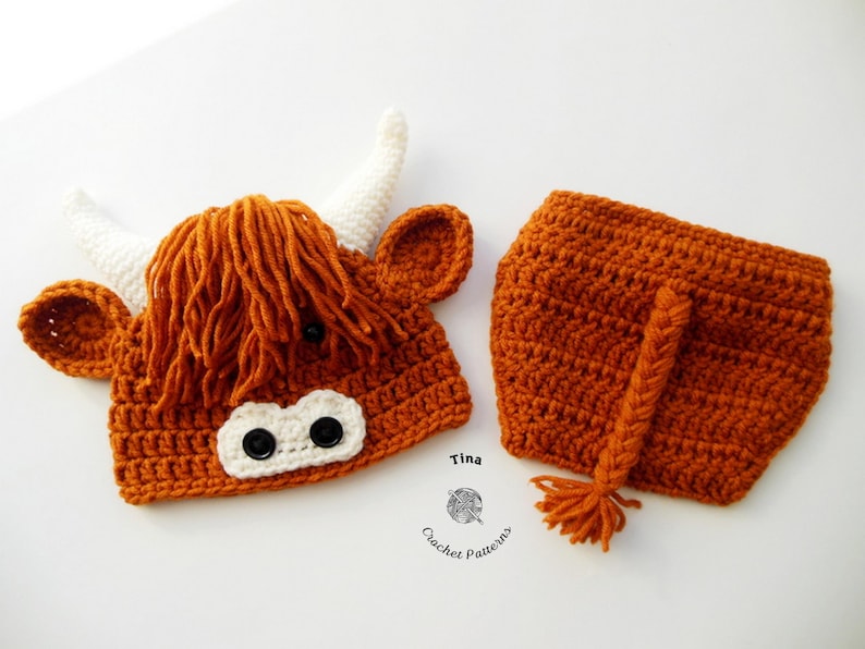 May include: A crocheted Highland cow costume set in shades of orange and white. The set includes a hat with horns, ears, and shaggy fringe, along with matching shorts with a tail. The hat features black button eyes and a white muzzle.