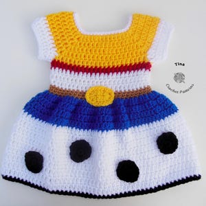 May include: A handmade crochet dress in yellow, white, blue, and red. The dress features a yellow bodice with white sleeves, a white midsection with a brown belt, and a blue skirt with black polka dots. The dress is a children's garment.