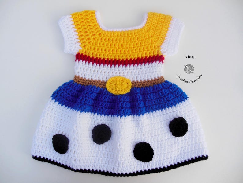 May include: A handmade crochet dress in yellow, white, blue, and red. The dress features a yellow bodice with white sleeves, a white midsection with a brown belt, and a blue skirt with black polka dots. The dress is a children's garment.