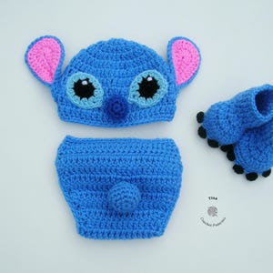 May include: A blue crocheted baby outfit with a Stitch character hat, diaper cover, and booties. The hat has pink ears, black eyes, and a blue nose. The booties have black pom-poms.