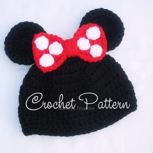 May include: A black crochet hat with two round ears and a red bow with white polka dots. The text "Crochet Pattern" is written in cursive on the hat. This handmade hat is a cute accessory.