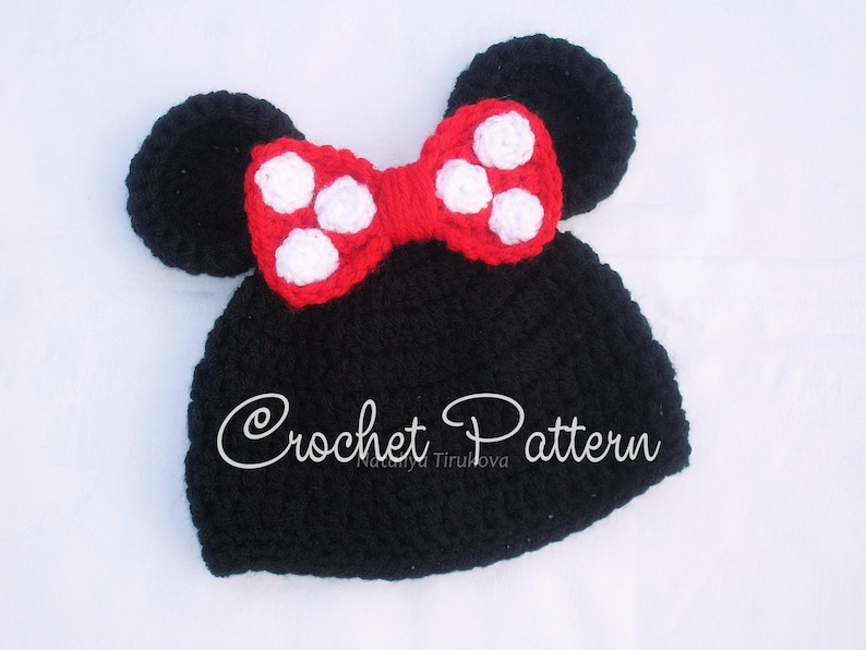 May include: A black crochet hat with two round ears and a red bow with white polka dots. The text "Crochet Pattern" is written in cursive on the hat. This handmade hat is a cute accessory.