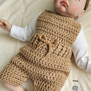 Op de afbeelding: Een bruine gebreide baby romper met een strikdetail. De romper is gemaakt met een eenvoudig steekpatroon en heeft een ontspannen pasvorm. De romper wordt gedragen door een babypop die op een wit deken ligt. De afbeelding bevat de tekst "Tina Crochet Patterns".