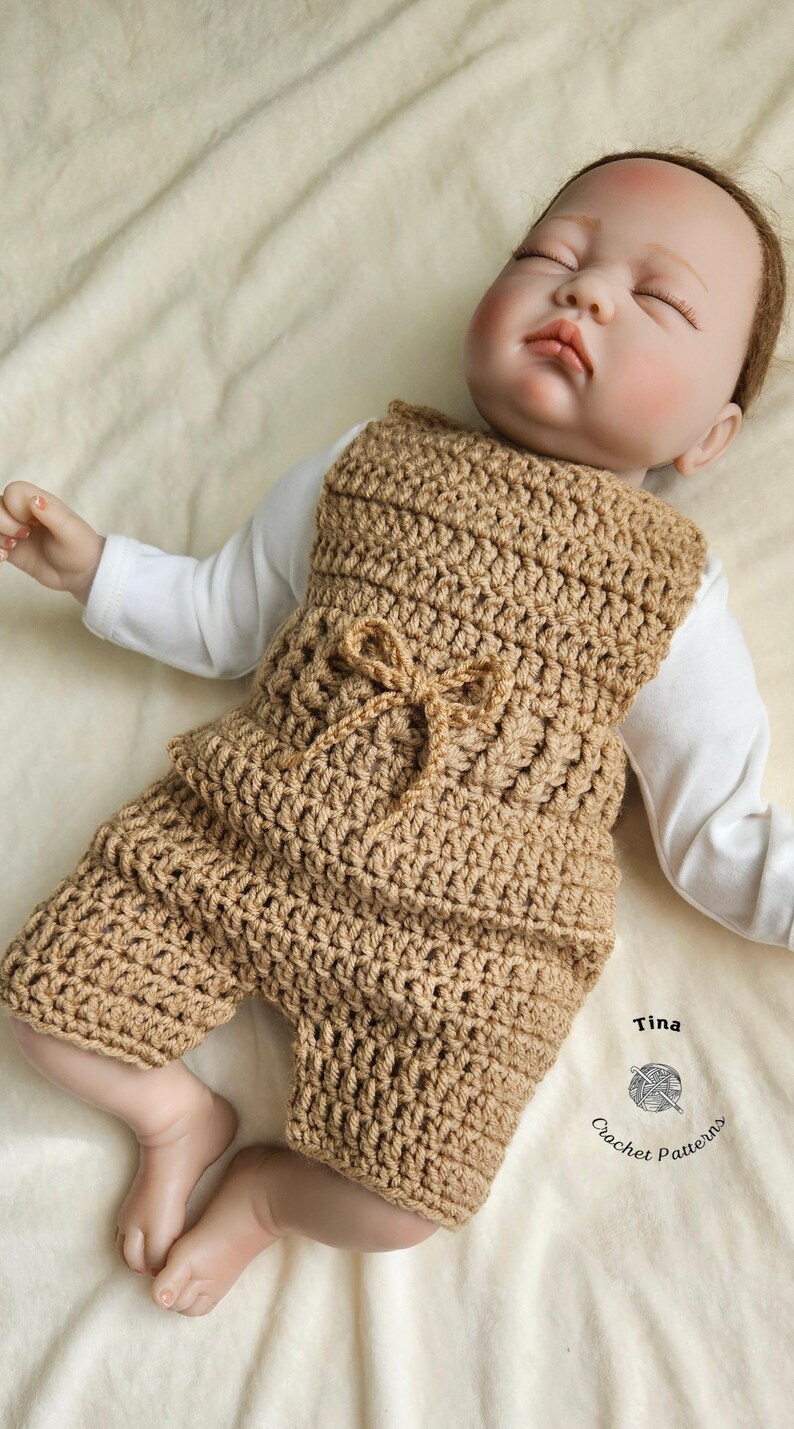Op de afbeelding: Een bruine gebreide baby romper met een strikdetail. De romper is gemaakt met een eenvoudig steekpatroon en heeft een ontspannen pasvorm. De romper wordt gedragen door een babypop die op een wit deken ligt. De afbeelding bevat de tekst "Tina Crochet Patterns".