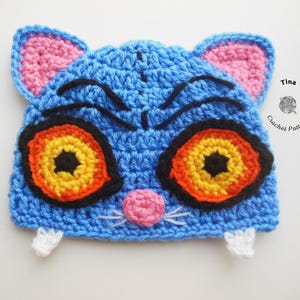 May include: A handmade blue crochet hat shaped like a cat's face. The hat features pink ears, large orange and yellow eyes with black outlines, a pink nose, white whiskers, and small white paws. The hat is a unique, whimsical accessory.