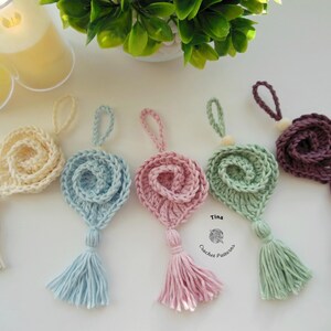 May include: Five crocheted flower ornaments with tassels in various colors, including cream, light blue, pink, green, and purple. The ornaments are hanging from a white surface. The text "Tina Crochet Patterns" is visible in the bottom right corner of the image.