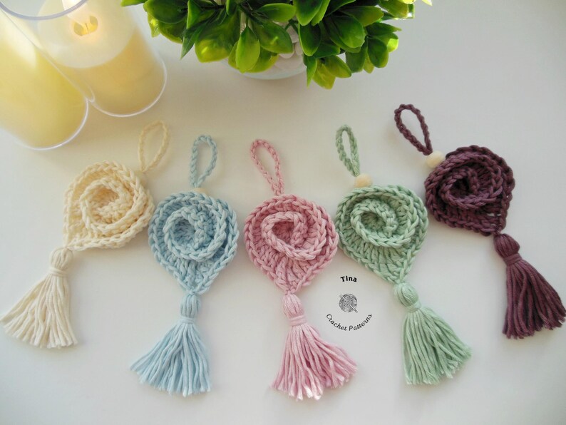 May include: Five crocheted flower ornaments with tassels in various colors, including cream, light blue, pink, green, and purple. The ornaments are hanging from a white surface. The text "Tina Crochet Patterns" is visible in the bottom right corner of the image.