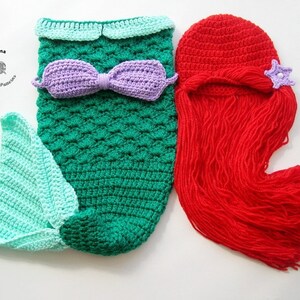 May include: A crocheted mermaid tail costume for a baby, with a green and blue tail, a purple bow, and a matching red crocheted wig with a purple star.