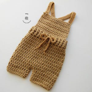 Op de afbeelding: Een gehaakte baby romper in een lichtbruine kleur met verstelbare bandjes en een strik in de taille.