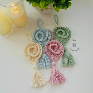 May include: Four crocheted flower ornaments with tassels in pastel colors: light blue, cream, pink, and green. The ornaments are hanging from a white surface. Tina Crochet Patterns is written on the surface.