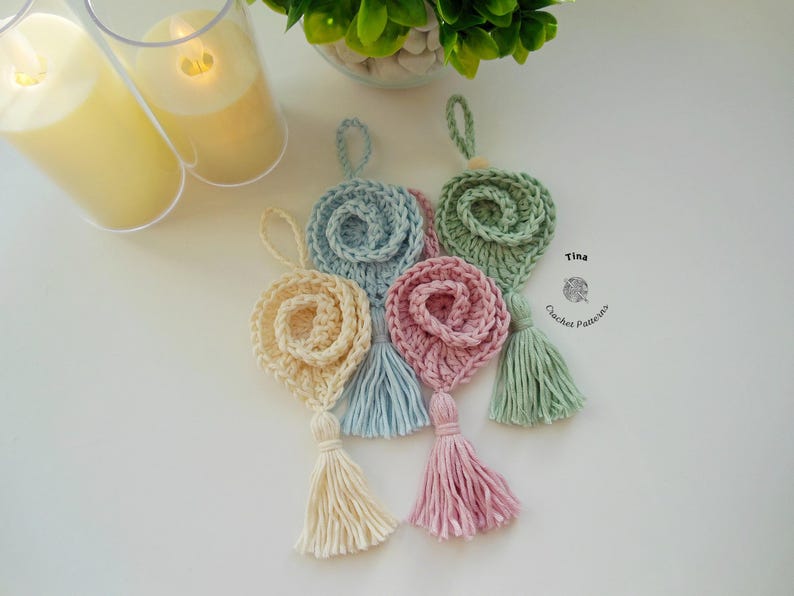 May include: Four crocheted flower ornaments with tassels in pastel colors: light blue, cream, pink, and green. The ornaments are hanging from a white surface. Tina Crochet Patterns is written on the surface.