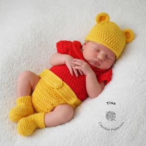 May include: A newborn baby wearing a handmade crochet outfit in yellow and red, resembling a bear. The outfit includes a hat with bear ears, a top, shorts, and booties. The baby is lying on a white, textured surface.