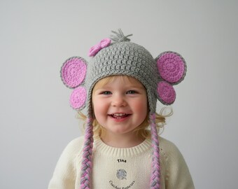 May include: A gray crochet elephant hat with pink ears and a matching bow. The hat has braided ear flaps in gray and pink. The child is wearing a cream-colored sweater. The text "Tina" and "Crochet Patterns" are visible.
