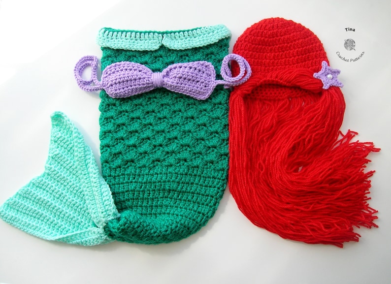 May include: A crocheted mermaid costume for a baby, featuring a green tail with a light blue fin, a purple top with a bow, and a red crocheted wig with a purple starfish.
