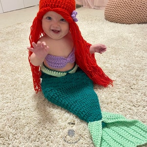May include: A baby dressed as a mermaid with a red crocheted hat and a purple crocheted top. The mermaid tail is green and light green. The baby is sitting on a white fluffy rug.
