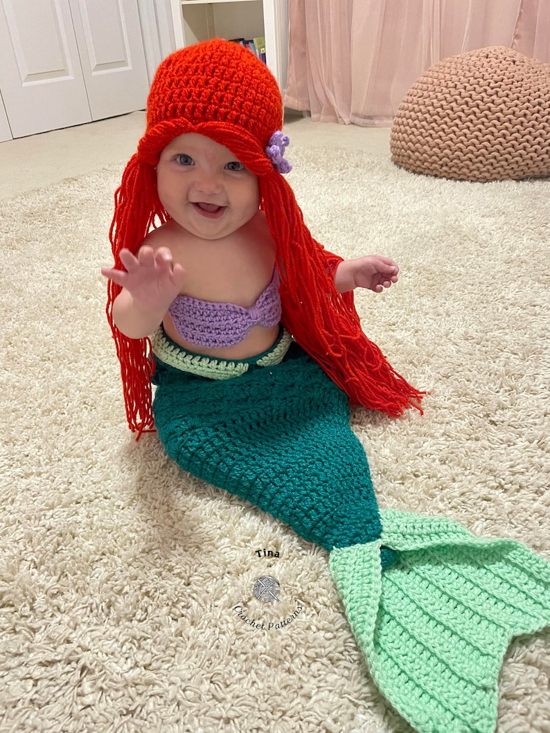 May include: A baby dressed as a mermaid with a red crocheted hat and a purple crocheted top. The mermaid tail is green and light green. The baby is sitting on a white fluffy rug.