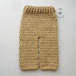 Può includere: Un paio di pantaloni beige all'uncinetto con una cintura larga. I pantaloni sono realizzati con un semplice motivo a punto e hanno una vestibilità rilassata.