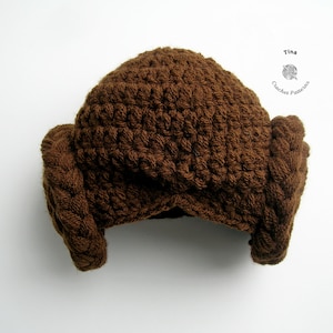 May include: A brown crocheted hat with ear flaps, resembling Princess Leia's hairstyle from Star Wars. The hat is made with a textured stitch and has a simple, classic design.