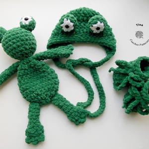 Könnte beinhalten: Grünes Häkel-Frosch-Plüschtier mit weißen Augen, eine passende grüne Häkelmütze mit weißen Augen und ein Paar grüne Häkel-Schühchen. Die Mütze hat eine lange Schleife.