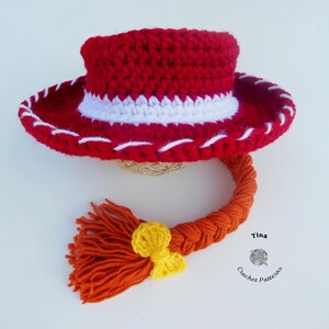 May include: A red crochet cowboy hat with a white band and stitching, and an orange braid with a yellow bow. The hat is handmade and likely for a child or baby. The hat is a costume or toy.