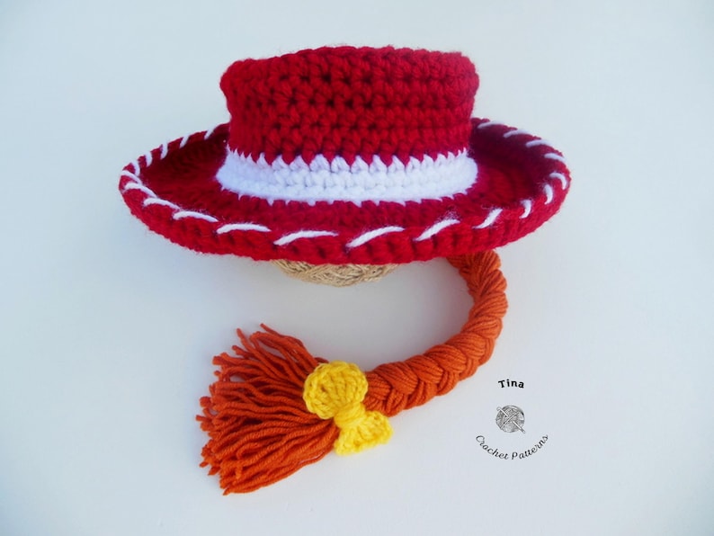 May include: A red crochet cowboy hat with a white band and stitching, and an orange braid with a yellow bow. The hat is handmade and likely for a child or baby. The hat is a costume or toy.