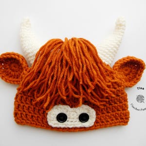 May include: A handmade crochet hat designed to resemble a Highland cow. The hat is primarily orange with white horns and ear details. The face features two black button eyes and a white patch.