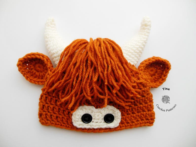 May include: A handmade crochet hat designed to resemble a Highland cow. The hat is primarily orange with white horns and ear details. The face features two black button eyes and a white patch.