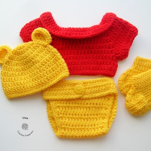 May include: A crocheted baby outfit in yellow and red. The outfit includes a hat, a diaper cover, a shirt, and booties. The hat has bear ears. The shirt has a red collar. The outfit is made with a yellow yarn.