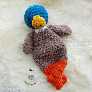 May include: A crocheted plush toy duck with a blue head, brown body, white beak, yellow beak detail, and orange feet. The duck is lying on a white fluffy surface. The text "Tina Crochet Patterns" is visible in the lower right corner of the image.