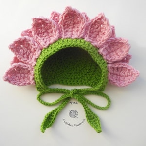 May include: A handmade crochet baby bonnet in the shape of a flower. The bonnet features pink petals, a green brim, and a green interior. The bonnet is tied with a green bow. The text "Tina Crochet Patterns" is visible.